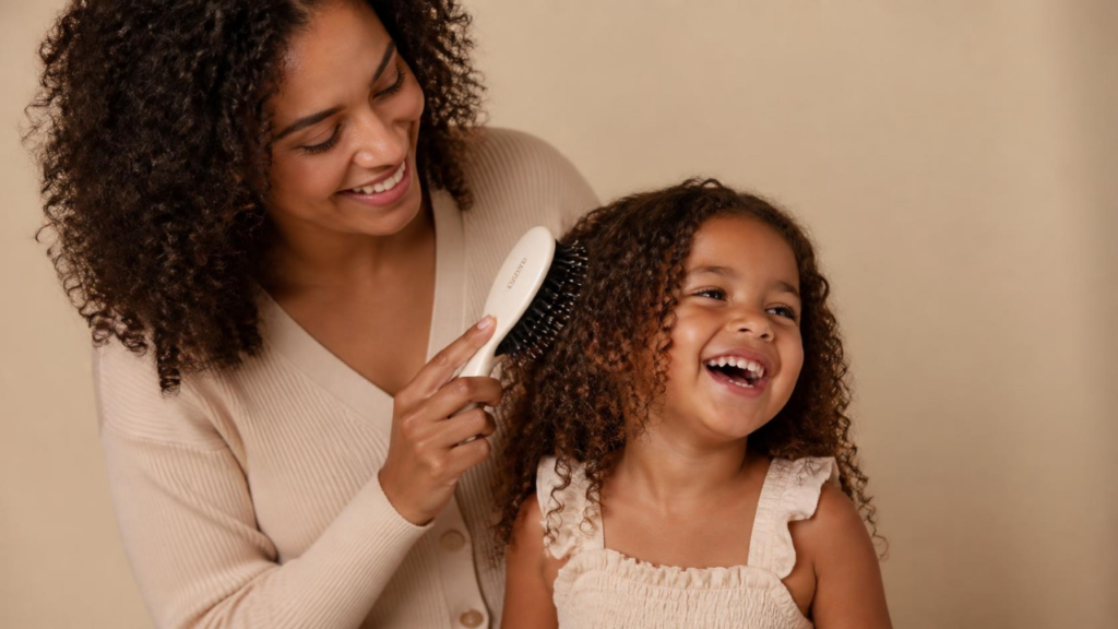 Uma mãe negra sorrindo enquanto usa uma escova suave para pentear o cabelo cacheado de sua filha pequena, que está rindo alegremente, ilustrando uma rotina capilar infantil sem dor.